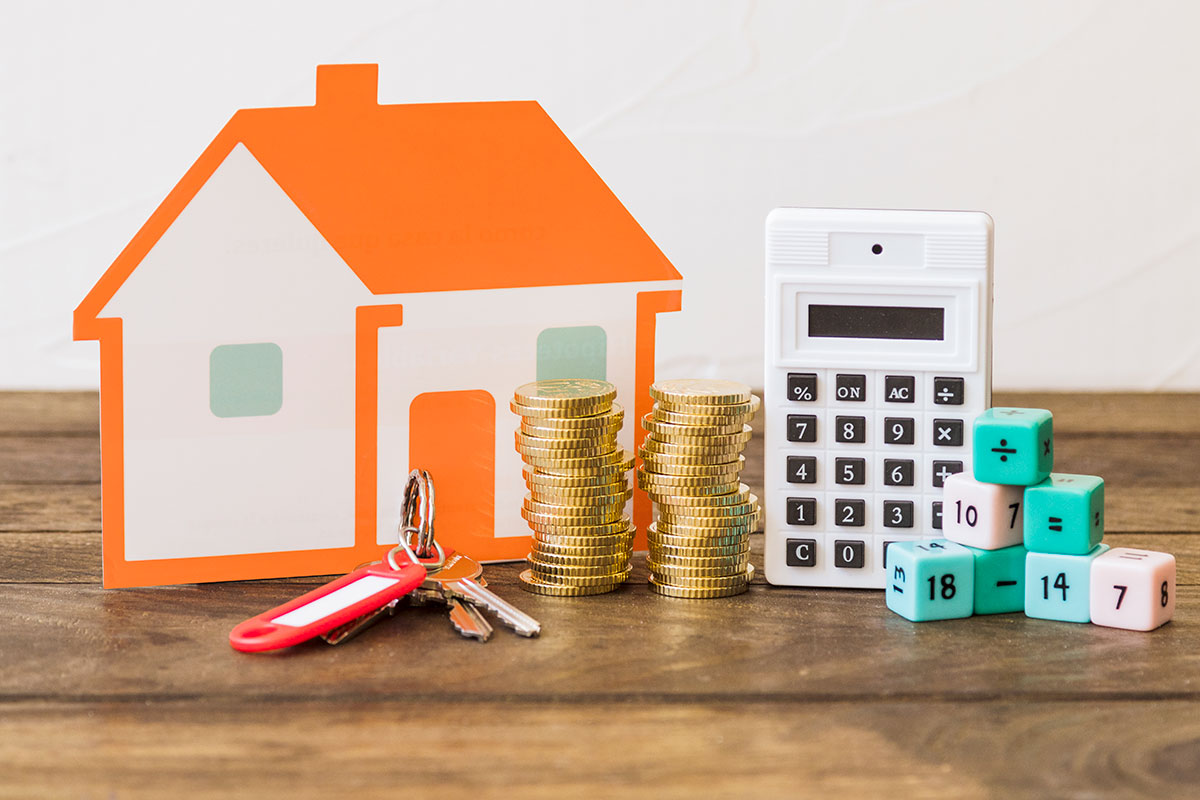 Imagen representativa del avalúo de casa, con una calculadora y monedas al lado de una casa de cartón. 
