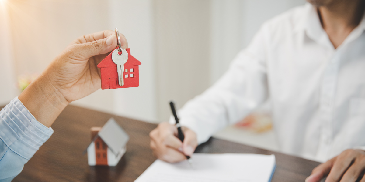 Foto ilustrativa de la venta de casas y terrenos, donde una mano sostiene unas llaves con un llavero en forma de casa, mientras se firma un contrato de compra de terreno.